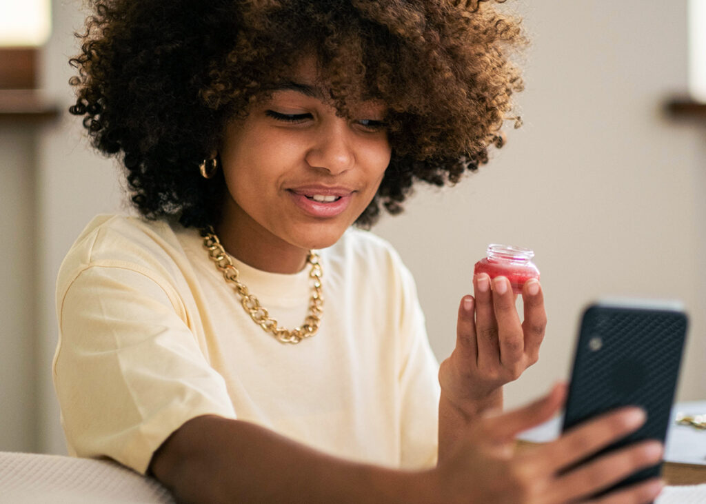 A brown skin curly haired woman in a white shirt with gold jewelry holding up a small product in front of her phone.
